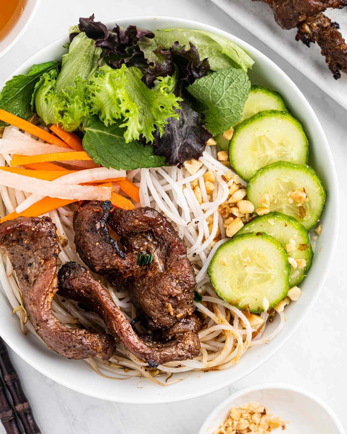 close up of bowl of bun thit nuong with vermicelli noodles, pickled carrots and daikon, assorted herbs and vegetables, grilled pork. Topped with crushed peanuts.